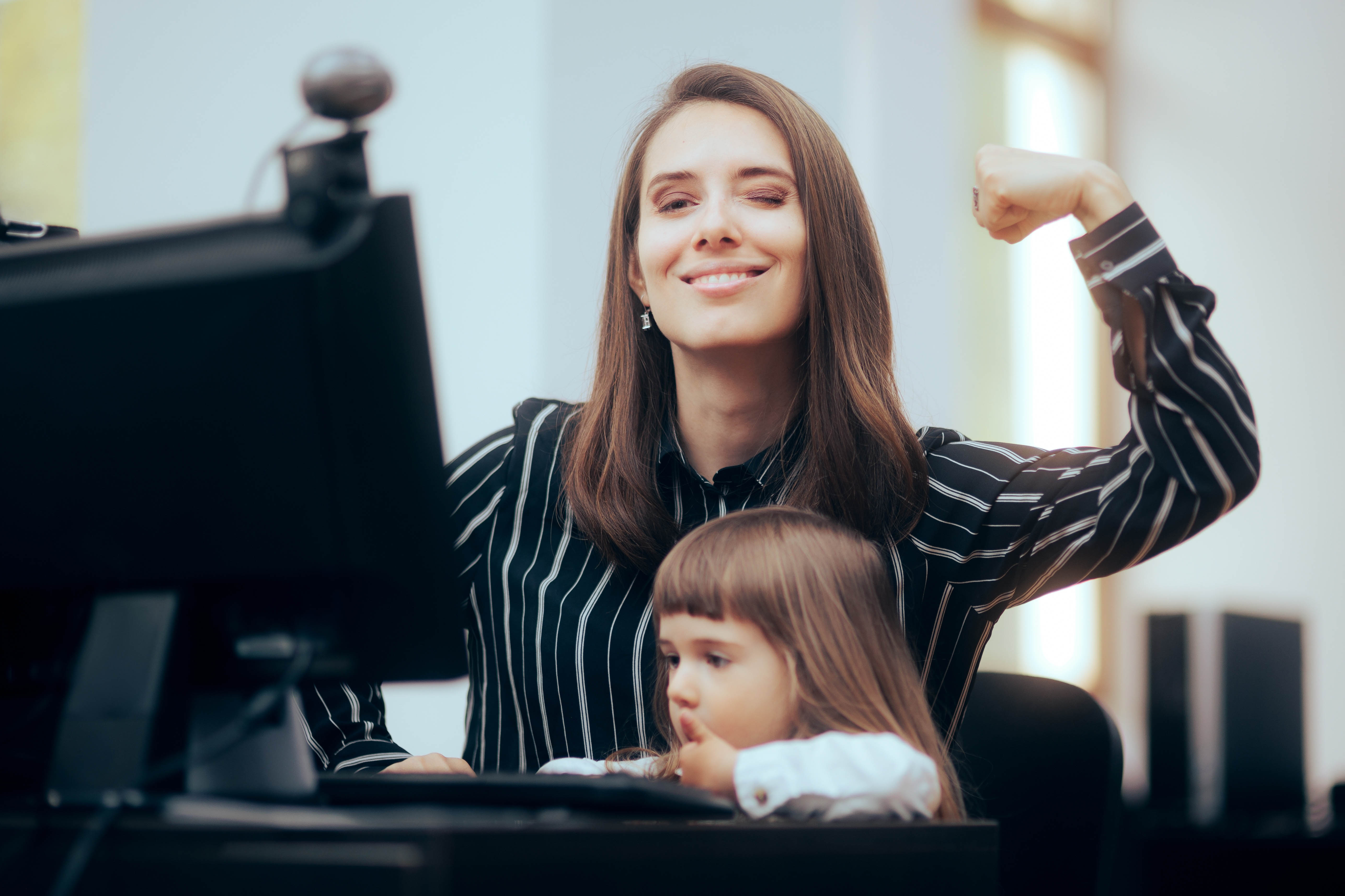 Professional looking young woman poses looking at the camera with her arm flexed, while a female toddler sits on her lap playing with the computer